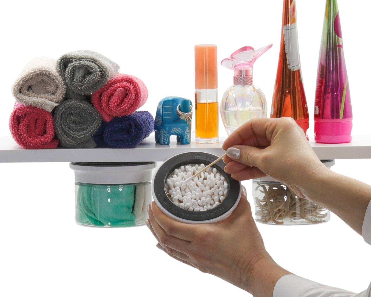 Hand accessing Q tip from white-lidded Cotton Ball Holder & Q Tip Organizer mounted under shelf with colorful bathroom decor.