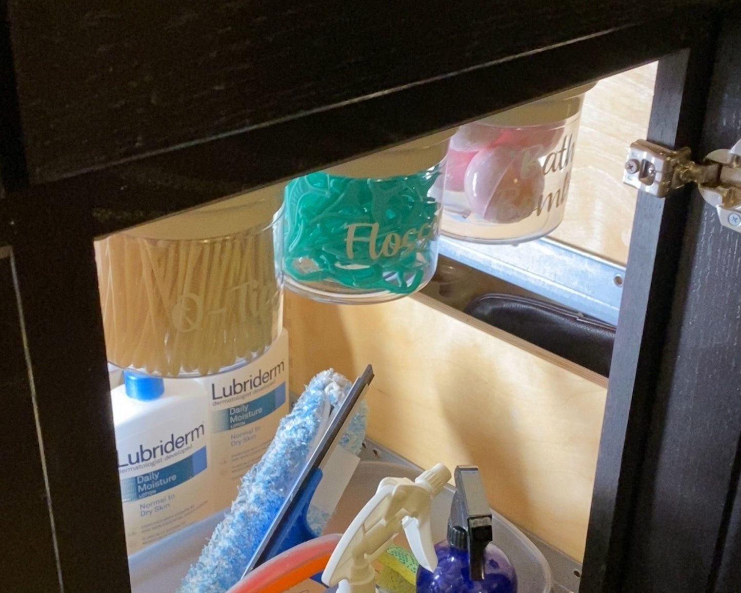 Clear labeled canisters for Q-Tips, Flossers, and Rounds mounted under bathroom sink. Efficient storage for small items.