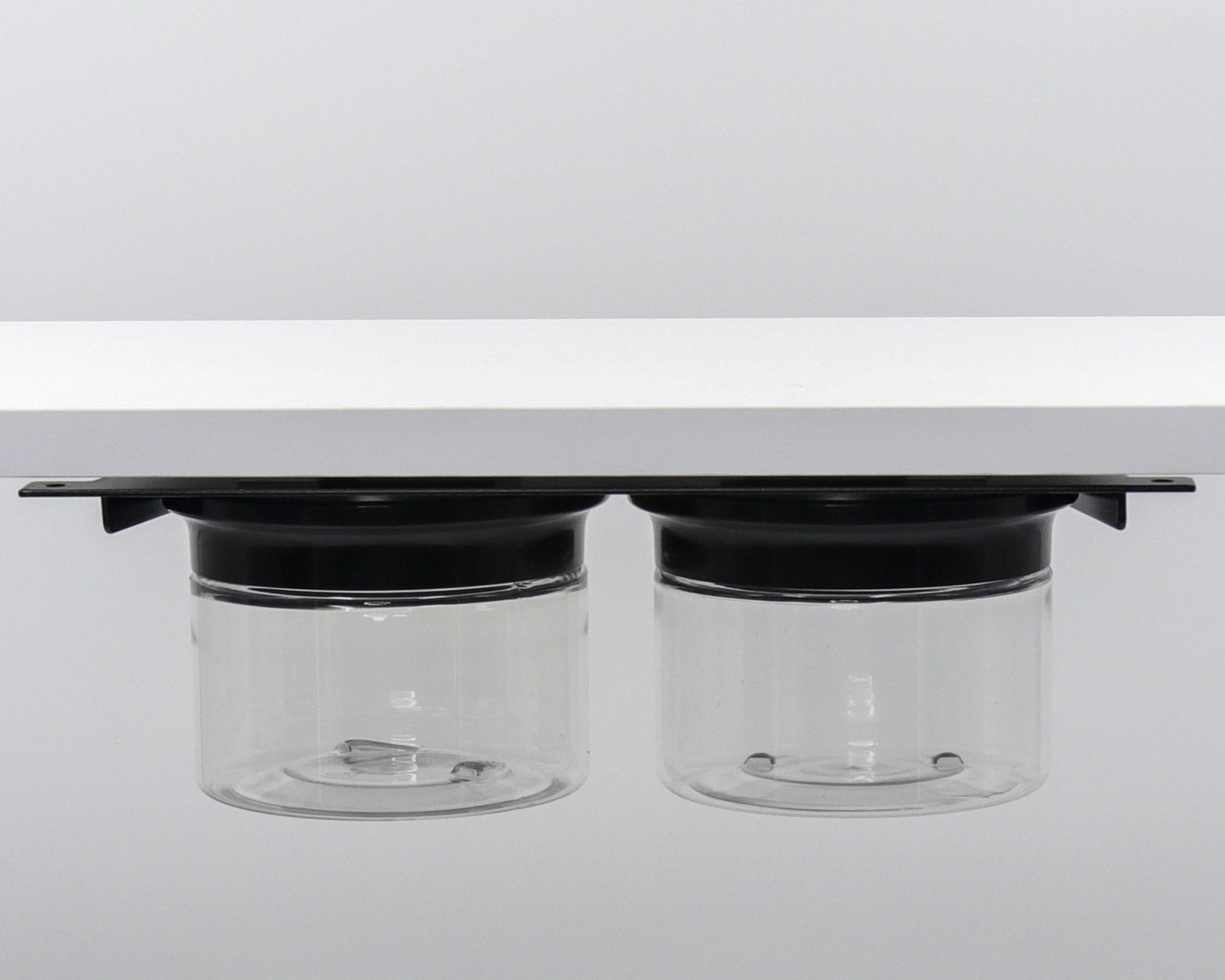 Two clear canisters with black lids mounted under shelf. Compact bathroom storage for cotton balls and flossers.