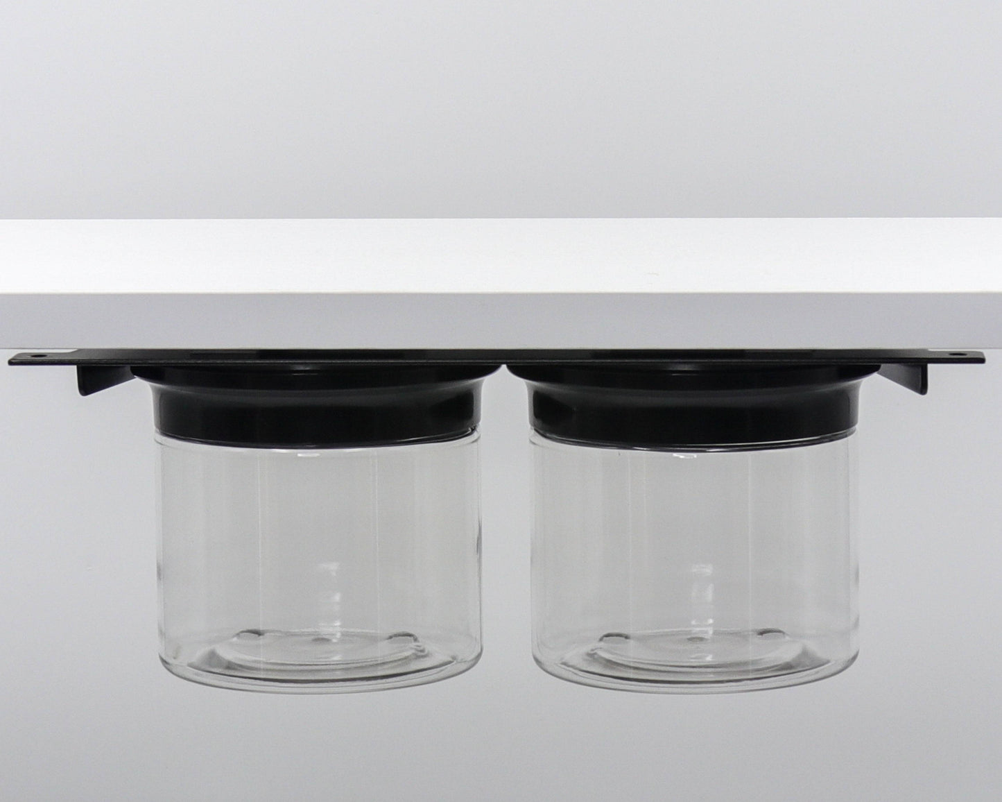 Dual canister set with black lids mounted under shelf. Space-saving bathroom organizer for small essentials.