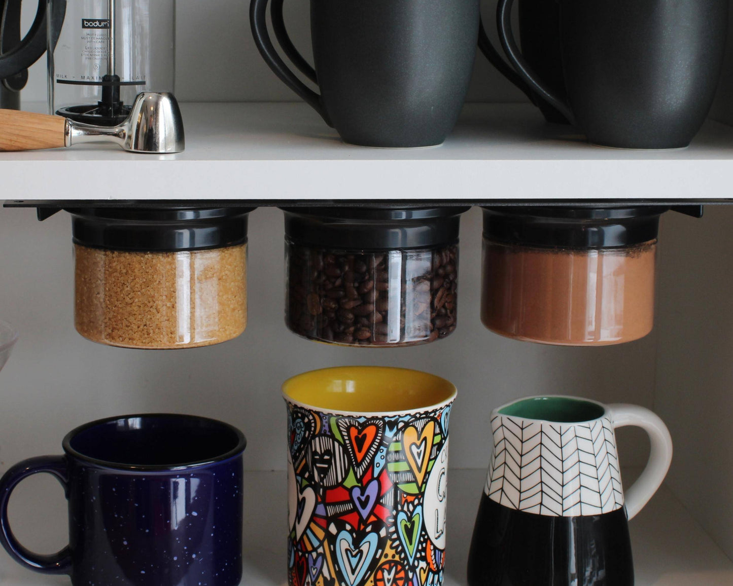 Under cabinet storage organizer with three airtight jars holding sugar, coffee beans, and cocoa powder. Space-saving kitchen solution for mugs and spices.