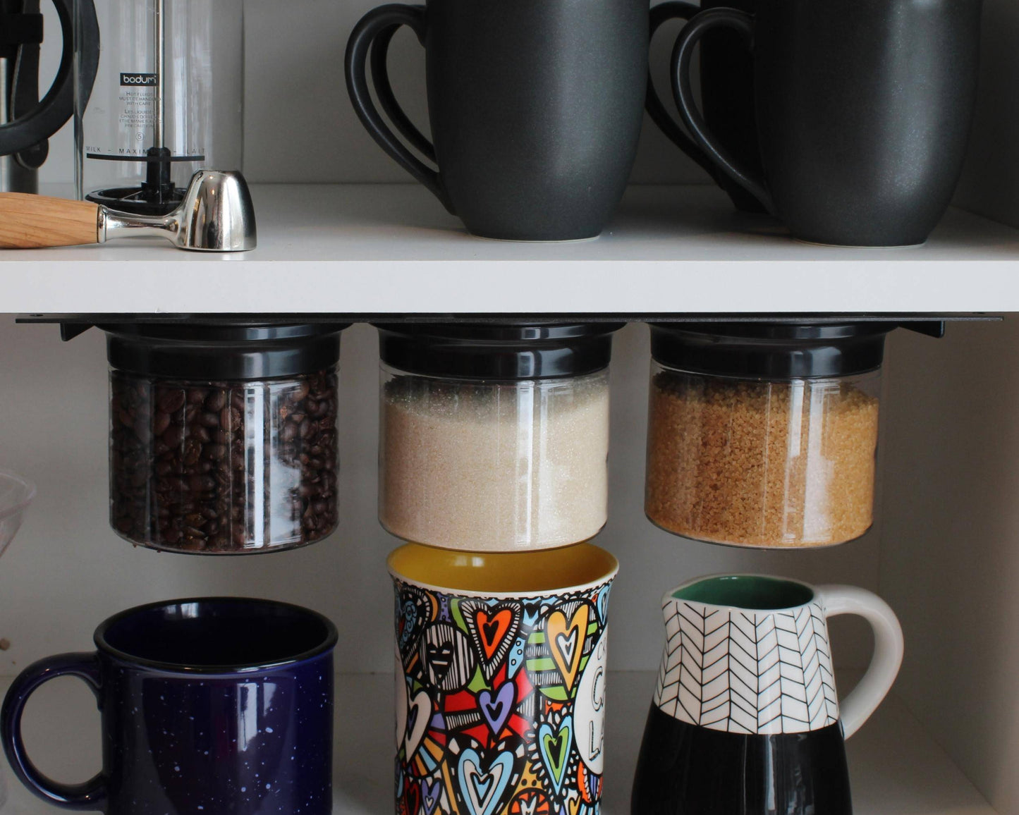 Under cabinet storage organizer with three airtight jars holding coffee beans, sugar, and brown sugar. Space-saving kitchen solution for mugs and spices.