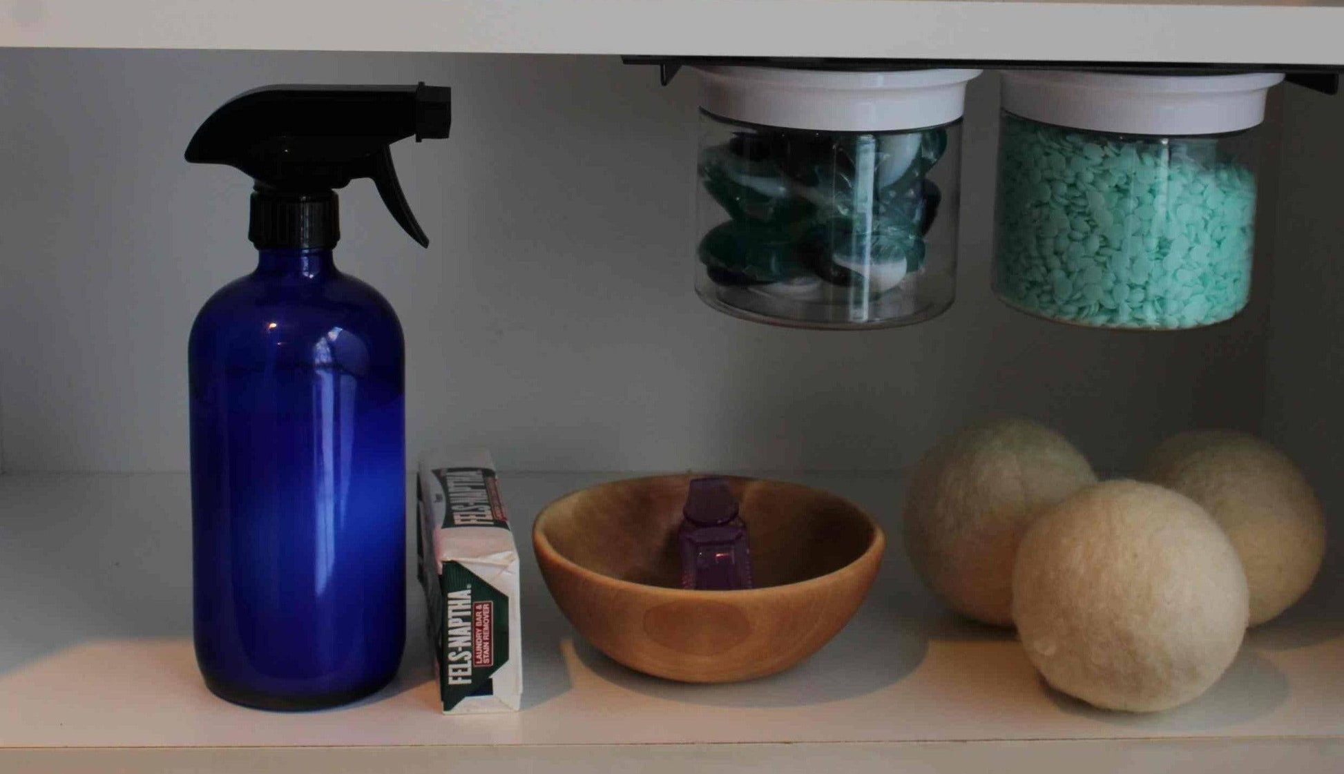 Under sink shelf with Laundry Containers Organizer jars storing detergent and scent beads, alongside laundry essentials like wool dryer balls and spray bottle.