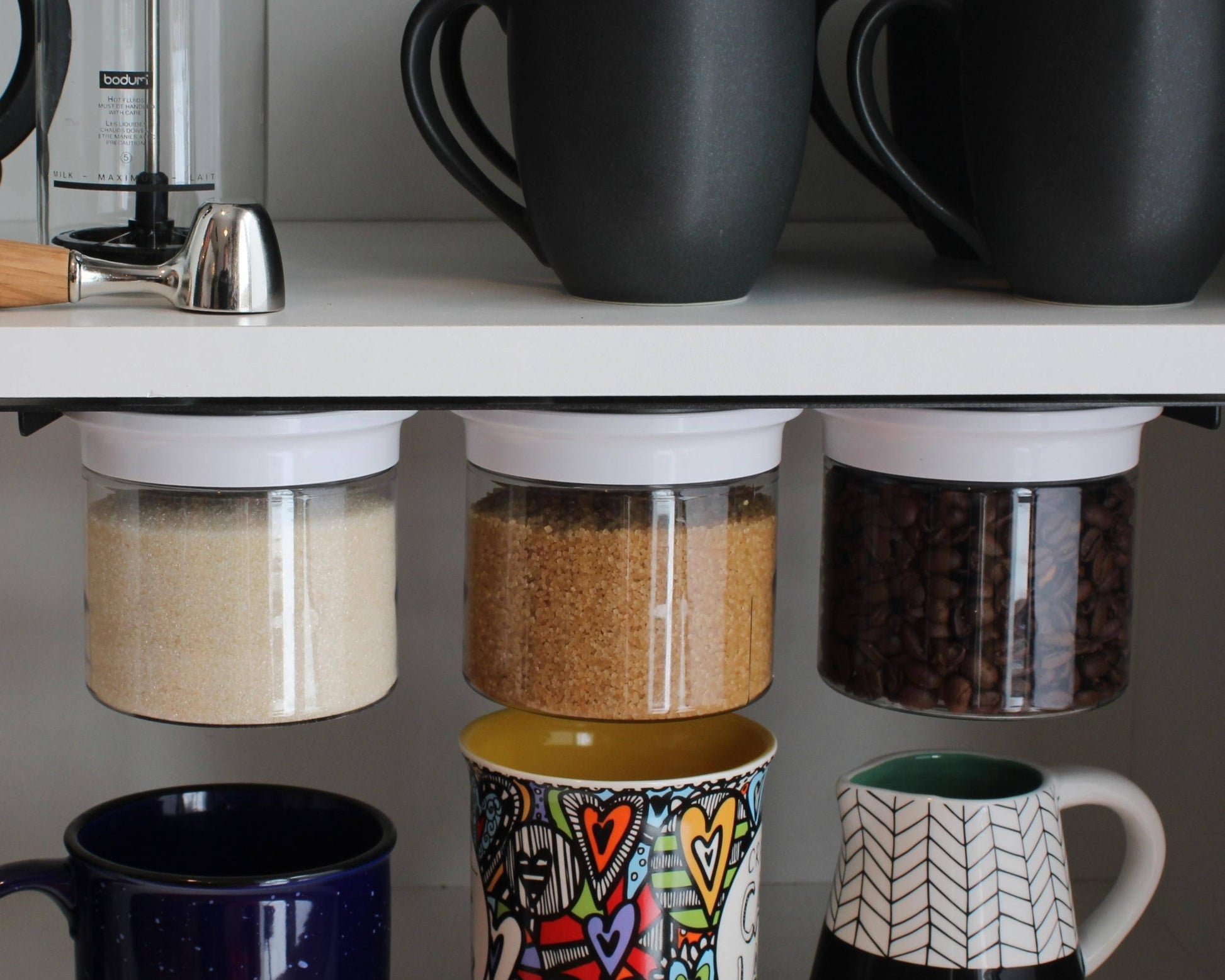 Under cabinet storage organizer with three airtight jars holding sugar, brown sugar, and coffee beans. Space-saving kitchen solution for mugs and spices.