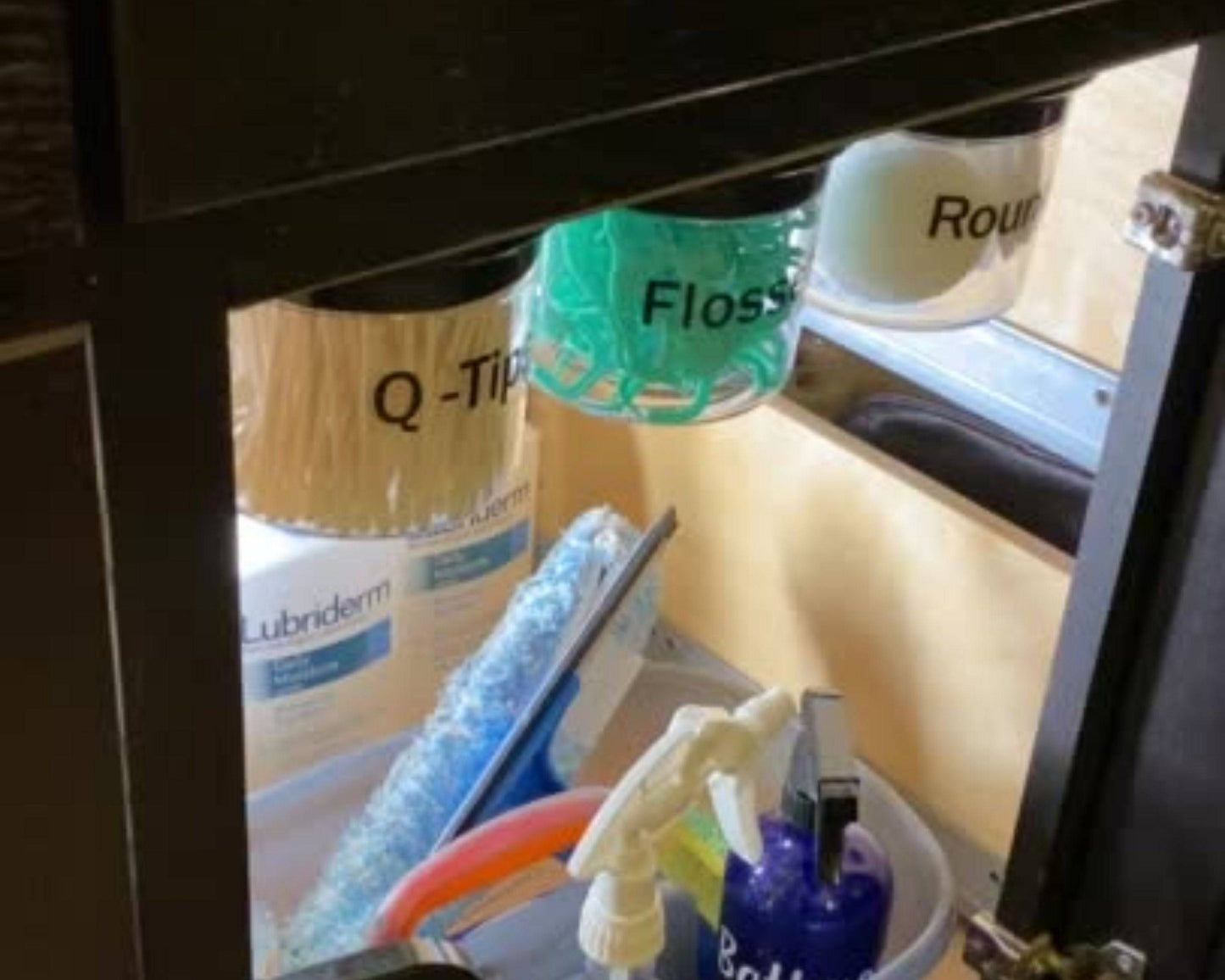 Mounted bathroom storage canisters under sink shelf labeled for Q-Tips, Flossers, and Rounds. Great for compact bathroom organization.