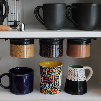 Airtight storage jars with magnetic rims mounted under a kitchen shelf, storing sugar, coffee beans, and cocoa powder above colorful mugs.