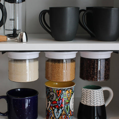 Three white-lid airtight jars mounted under a kitchen shelf, storing sugar, brown sugar, and coffee beans above colorful mugs.